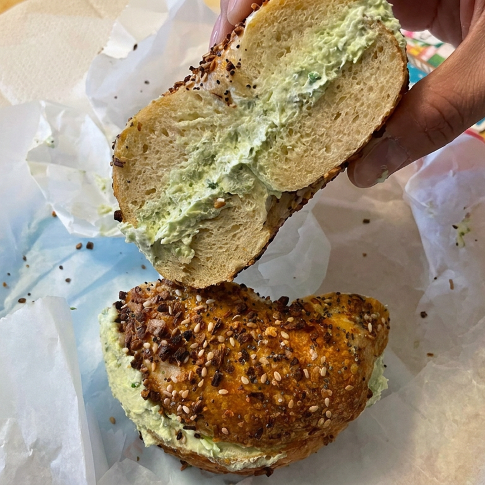 Hand lifts the top of a seeded bagel sandwich, revealing a thick layer of pale green spread on wax paper.