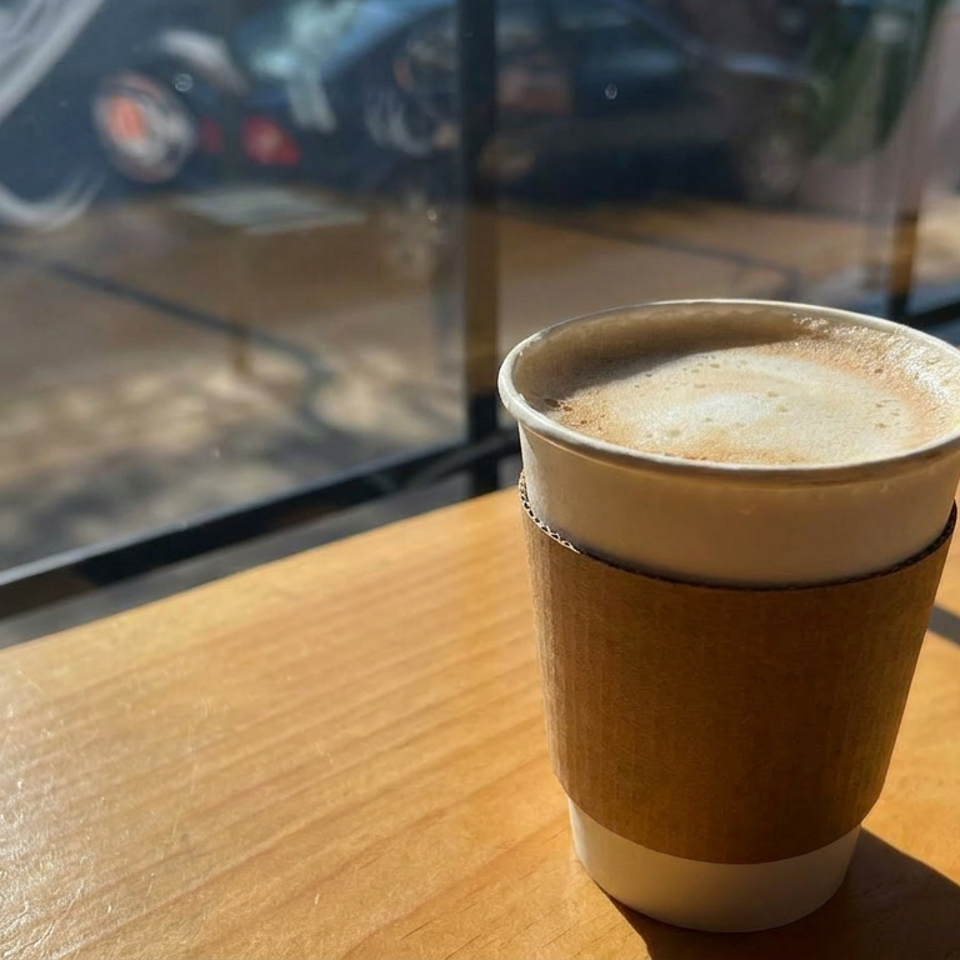 Foamy coffee in a paper cup sits on a sunlit wooden table beside a café window.